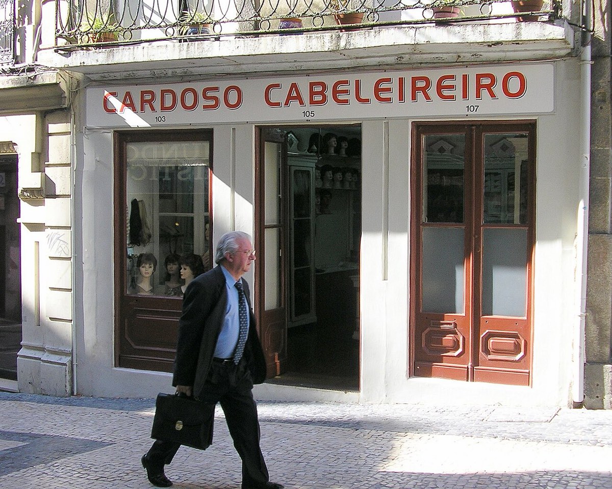 Fachada do Cardoso Cabeleireiro no Porto, com letreiro vermelho, portas de madeira e um homem de fato a passar no passeio