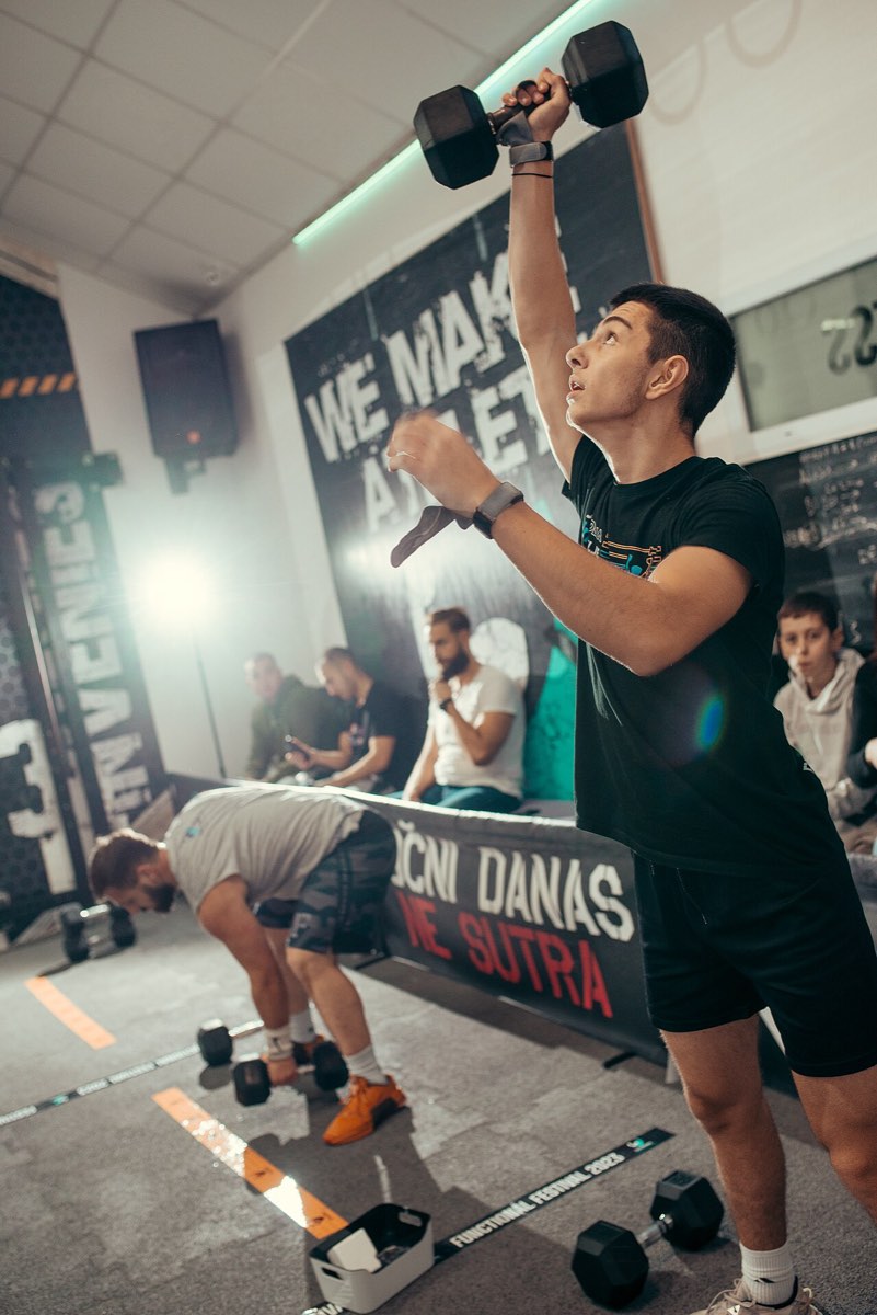 Jovem de t-shirt preta a levantar um haltere acima da cabeça numa aula de treino funcional, com outro atleta e público ao fundo